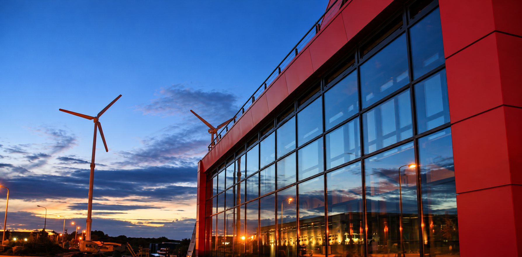 Façade vitrée d'un bâtiment moderne aux panneaux rouges, avec deux éoliennes se détachant sur un ciel bleu au crépuscule.