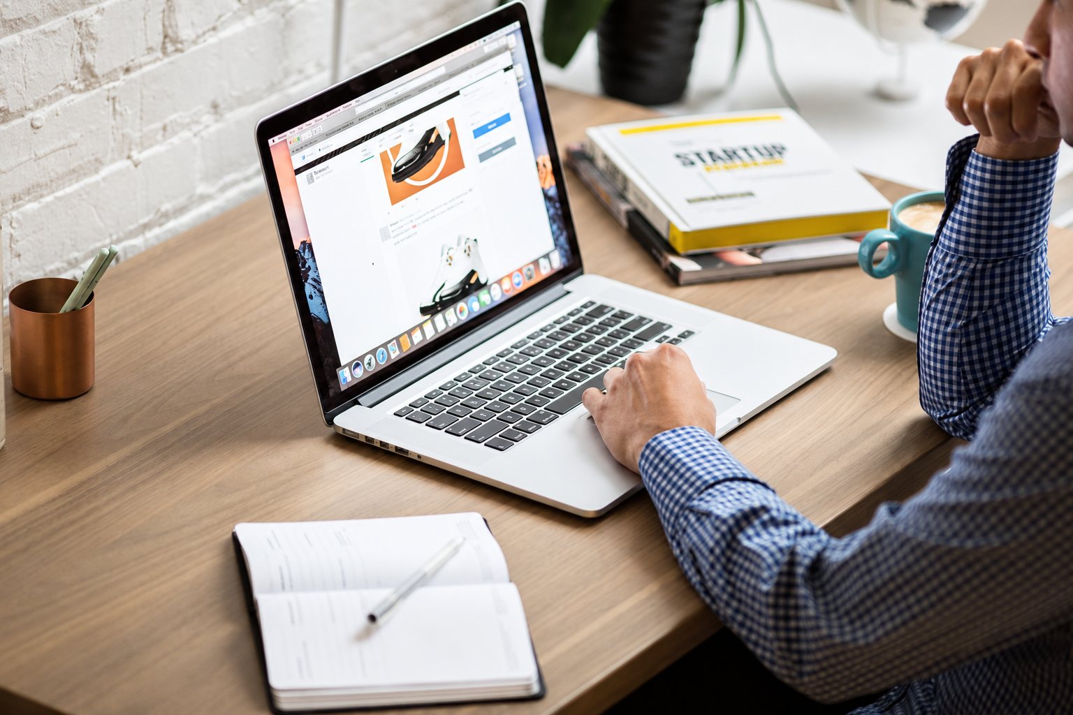 Un homme en chemise à carreaux utilise un ordinateur portable posé sur un bureau en bois, à côté d'un carnet ouvert, d'une tasse et de livres.
