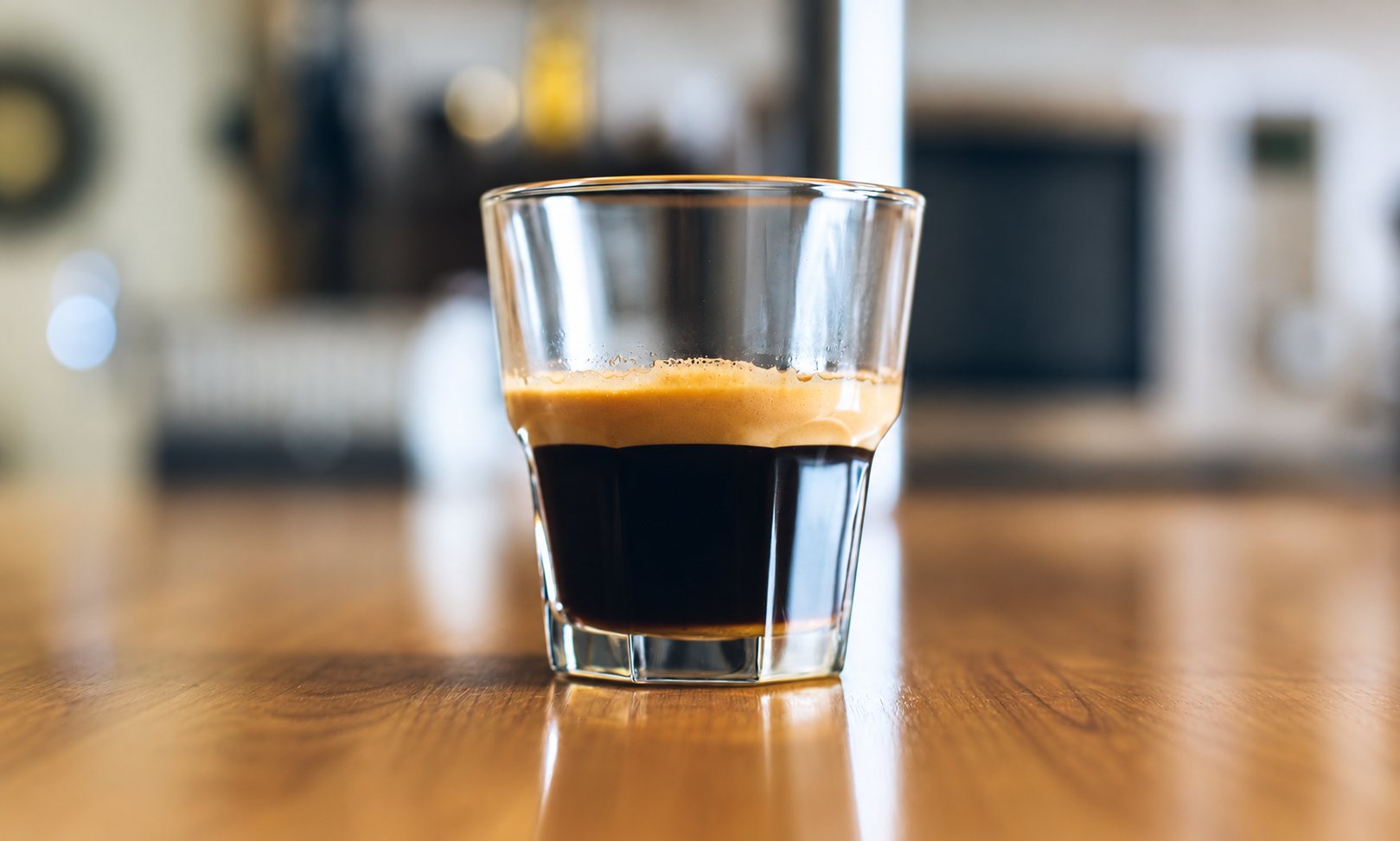 Un espresso avec une belle crème servi dans un petit verre transparent posé sur une table en bois, devant un arrière-plan flou.